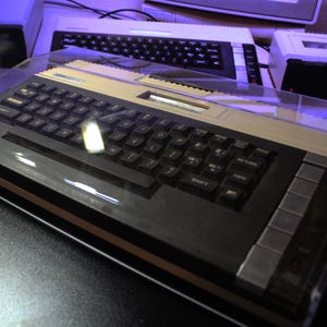 May include: A vintage computer with a black keyboard and beige accents, encased in a clear protective cover. The keyboard has numerous keys, and the design suggests a retro aesthetic. Other computer components are visible in the background.