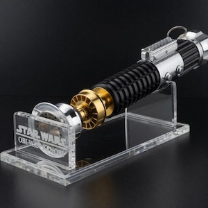 May include: A Star Wars Obi-Wan Kenobi lightsaber replica, featuring a black, silver, and gold design. The lightsaber is displayed on a clear acrylic stand with the Star Wars logo and character name. The lightsaber has a detailed handle and a metallic emitter.