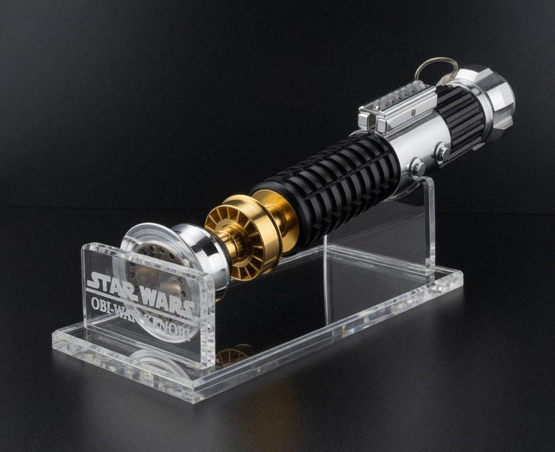 May include: A Star Wars Obi-Wan Kenobi lightsaber replica, featuring a black, silver, and gold design. The lightsaber is displayed on a clear acrylic stand with the Star Wars logo and character name. The lightsaber has a detailed handle and a metallic emitter.