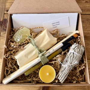 May include: A kraft paper box filled with art supplies and spiritual items. Includes a sage bundle, paintbrushes, pencils, a candle, a crystal, and a card with the text "Quiet mind. Open heart. Create."