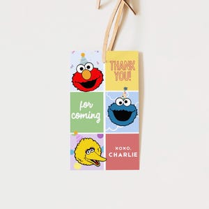 May include: A colorful gift tag featuring Sesame Street characters Elmo, Cookie Monster, and Big Bird. The tag includes the phrases "Thank You!" and "for coming", along with "XOXO, Charlie". The tag is attached to a wooden string.