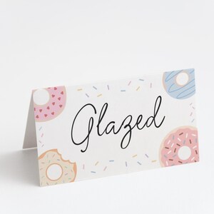 May include: A folded card with the word "Glazed" in cursive script, surrounded by illustrations of donuts with pink, blue, and tan frosting, and colorful sprinkles. The card is on a white background.