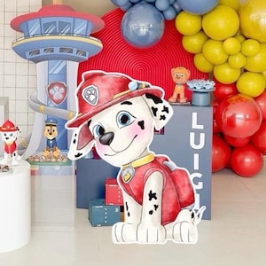 Op de afbeelding: Een grote cartoon uitsparing van Marshall van Paw Patrol, met een rode brandweerhoed en vest. De achtergrond bevat een Paw Patrol toren, ballonnen en karakter beeldjes, wat wijst op een feest thema.