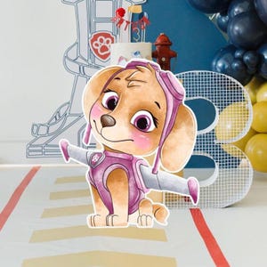 Puede incluir: Un recorte de dibujos animados de Skye de Paw Patrol, con un uniforme rosa y blanco con alas. El fondo incluye una tarta de cumpleaños, globos y una boca de incendios. El personaje sonríe con mejillas rosadas.