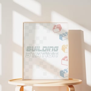 May include: A framed print with the words "BUILDING STATION" in a stylized font. The background is a light gray checkered pattern. Colorful toy brick illustrations are scattered around the frame.