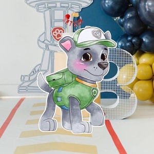 May include: A cutout of the cartoon character Rocky from Paw Patrol. Rocky is a gray dog wearing a green vest, backpack, and cap. The background includes a large number 3, balloons, and a Paw Patrol tower.