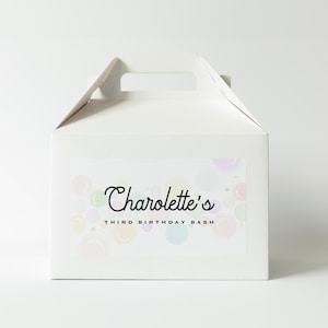 Peut inclure: Une boîte blanche à cadeaux avec une poignée, ornée d'une étiquette avec le texte "Charolette's THIRD BIRTHDAY BASH" en écriture noire. L'étiquette présente un motif de bulles aux couleurs pastel.