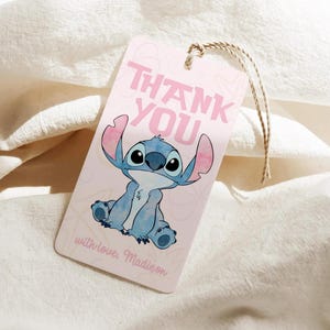 Puede incluir: Una etiqueta de regalo rosa con una ilustraci&oacute;n de un personaje de dibujos animados azul y las palabras "THANK YOU" en rosa. La etiqueta tambi&eacute;n incluye el texto "with love, Madison" en cursiva y est&aacute; sujeta a una cuerda de hilo natural.