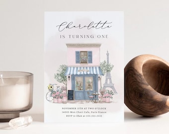 May include: A watercolor invitation with the text "Charolette is turning one." The design features a pink cafe with a blue awning, a bicycle, and the Eiffel Tower. Additional text includes the date, time, and RSVP information.