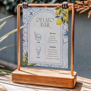 Puede incluir: Un letrero de bar de helados de madera con un marco de cobre. El letrero presenta un diseño de azulejos azules y blancos y enumera sabores de helado como limón y pistacho, y aderezos como rodajas de melocotón y frutos secos variados.