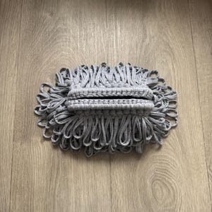 Può includere: Una pochette grigia in tessuto fatta a mano con un design a frange ad anello. La pochette è realizzata in un materiale morbido e lavorato a maglia e ha una forma rettangolare. La frangia è ad anello e crea un elemento decorativo strutturato.