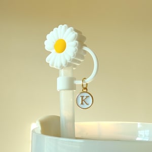 May include: White and yellow daisy-shaped bottle stopper with a gold-colored charm featuring the letter "K". The stopper is designed to fit into a bottle. The daisy has white petals and a yellow center.