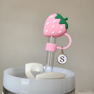 Può includere: Un tappo per cannuccia a forma di fragola rosa con dettagli di semi bianchi e un gambo verde. La cannuccia trasparente ha una fascia rosa e un ciondolo con la lettera "S". La cannuccia è attaccata a un bicchiere bianco e trasparente.