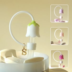 May include: A white and green flower-shaped straw topper with a gold initial charm. The topper is attached to a white curved straw guide. Additional images show similar toppers in pink and purple.