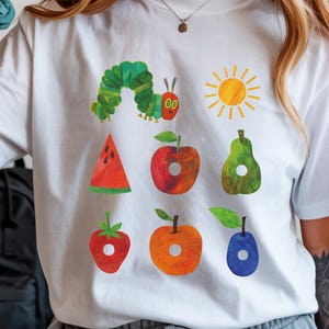 May include: White t-shirt featuring a colorful illustration of a green caterpillar, a yellow sun, and various fruits. The design includes a watermelon slice, apple, pear, strawberry, orange, and a plum. The word "WHITE" is printed at the top right.