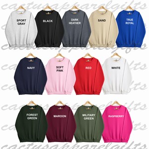Puede incluir: Un conjunto de 12 sudaderas de cuello redondo en varios colores, incluyendo gris deportivo, negro, gris oscuro jaspeado, arena, azul real, azul marino, rosa suave, rojo, blanco, verde bosque, granate, verde militar y frambuesa. Cada sudadera tiene un color diferente y una etiqueta de texto blanca con el nombre del color.