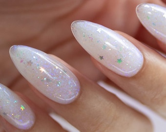 Puede incluir: Primer plano de una mano con uñas largas, almendradas e iridiscentes. Las uñas son de un blanco perlado con un sutil tono lavanda y están adornadas con purpurina brillante y pequeños detalles en forma de estrella. El efecto general es etéreo.