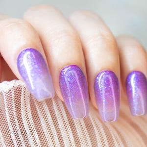 May include: Close-up of a hand with five long, oval-shaped nails painted with a gradient of light purple to dark purple glitter polish.