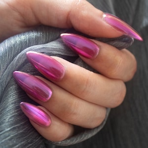 May include: A hand displaying long, almond-shaped nails painted in a metallic pink shade. The nails have a glossy, reflective surface that shimmers. The hand is holding a piece of grey fabric, providing a contrasting backdrop to the bright nail colour.
