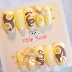 Chocolate banana inspired nails. Pastel yellow base with banana fruit slices, chocolate spread, and chocolate drizzle, with 3d stars and chocolate chunks. A cute brown ribbon adds a charming touch.