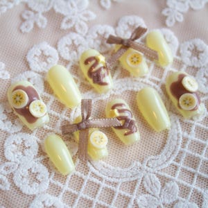 Chocolate banana inspired nails. Pastel yellow base with banana fruit slices, chocolate spread, and chocolate drizzle, with 3d stars and chocolate chunks. A cute brown ribbon adds a charming touch.