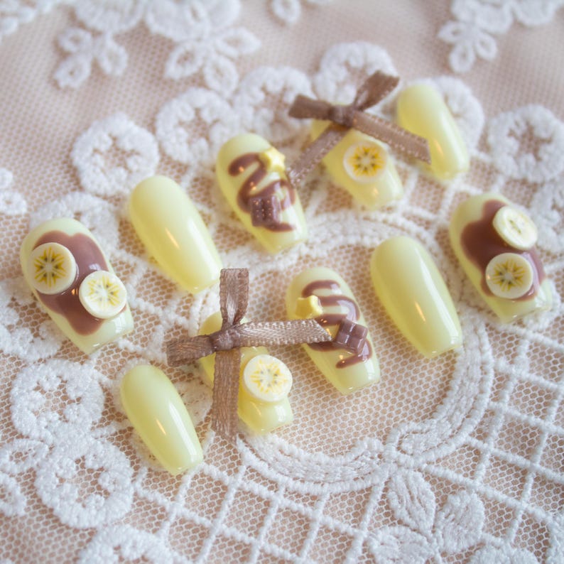 Chocolate banana inspired nails. Pastel yellow base with banana fruit slices, chocolate spread, and chocolate drizzle, with 3d stars and chocolate chunks. A cute brown ribbon adds a charming touch.