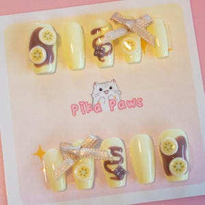 Chocolate banana inspired nails. Pastel yellow base with banana fruit slices, chocolate spread, and chocolate drizzle, with 3d stars and chocolate chunks. A cute brown ribbon adds a charming touch.