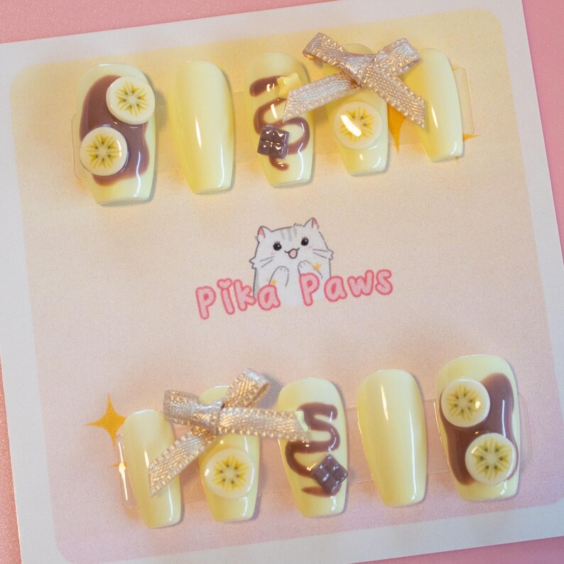 Chocolate banana inspired nails. Pastel yellow base with banana fruit slices, chocolate spread, and chocolate drizzle, with 3d stars and chocolate chunks. A cute brown ribbon adds a charming touch.