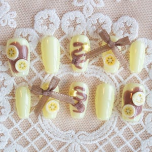 Chocolate banana inspired nails. Pastel yellow base with banana fruit slices, chocolate spread, and chocolate drizzle, with 3d stars and chocolate chunks. A cute brown ribbon adds a charming touch.