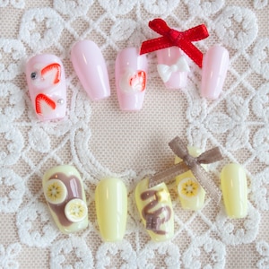 Chocolate banana inspired nails. Pastel yellow base with banana fruit slices, chocolate spread, and chocolate drizzle, with 3d stars and chocolate chunks. A cute brown ribbon adds a charming touch. Pictured is also the "Strawberry Darling" set.