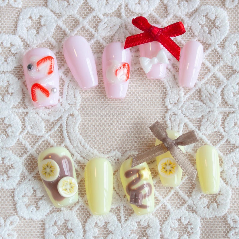 Chocolate banana inspired nails. Pastel yellow base with banana fruit slices, chocolate spread, and chocolate drizzle, with 3d stars and chocolate chunks. A cute brown ribbon adds a charming touch. Pictured is also the "Strawberry Darling" set.