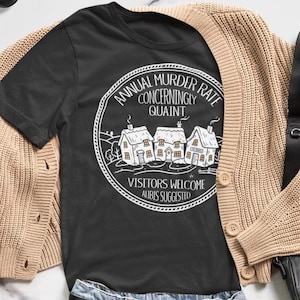 Op de afbeelding: Een zwart t-shirt met een witte cirkelvormige afbeelding van drie kleine huisjes en de tekst: ANNUAL MURDER RATE CONCERNINGLY QUAINT, VISITORS WELCOME, ALIBIS SUGGESTED. Het t-shirt wordt gedragen onder een beige gebreid vest met knopen.