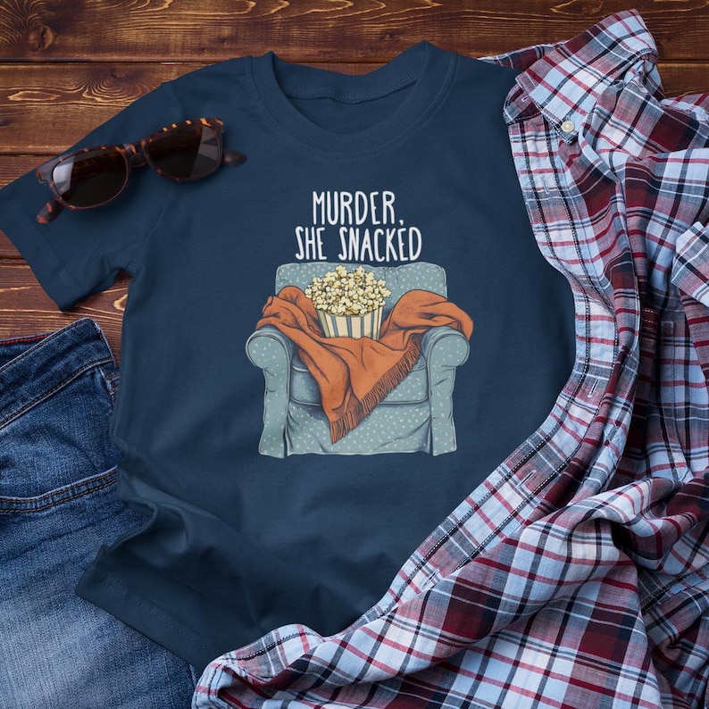Op de afbeelding: Een marineblauw t-shirt met de tekst "Murder, She Snacked" en een cartoonillustratie van een persoon die in een fauteuil zit met een kom popcorn.