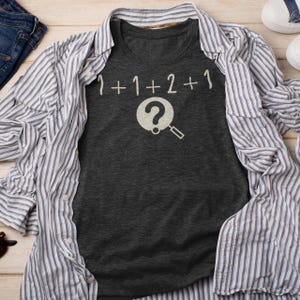 May include: A dark gray t-shirt with a white graphic of a magnifying glass over a question mark and the text "1+1+2+1".