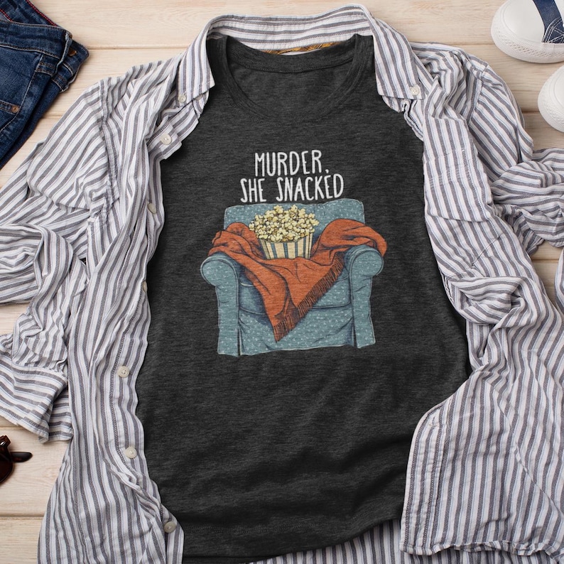 Op de afbeelding: Een donkergrijs T-shirt met een afbeelding van een persoon die in een stoel zit met een deken en een kom popcorn. De tekst "Murder, She Snacked" staat op het shirt gedrukt.