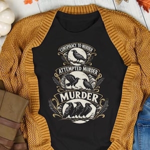 Op de afbeelding: Een zwart t-shirt met een grafisch ontwerp van drie lagen kraaien en tekst. De bovenste laag zegt CONSPIRACY TO MURDER met één kraai, het midden zegt ATTEMPTED MURDER met twee kraaien en de onderkant zegt MURDER met drie kraaien.
