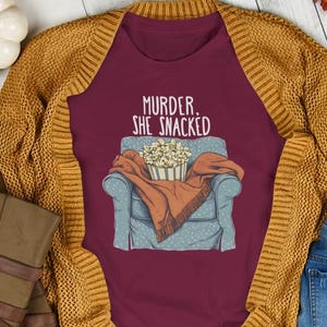 Op de afbeelding: Een maroon t-shirt met een afbeelding van iemand die in een fauteuil zit met een deken en een kom popcorn. De tekst "Murder, She Snacked" staat op het shirt gedrukt.