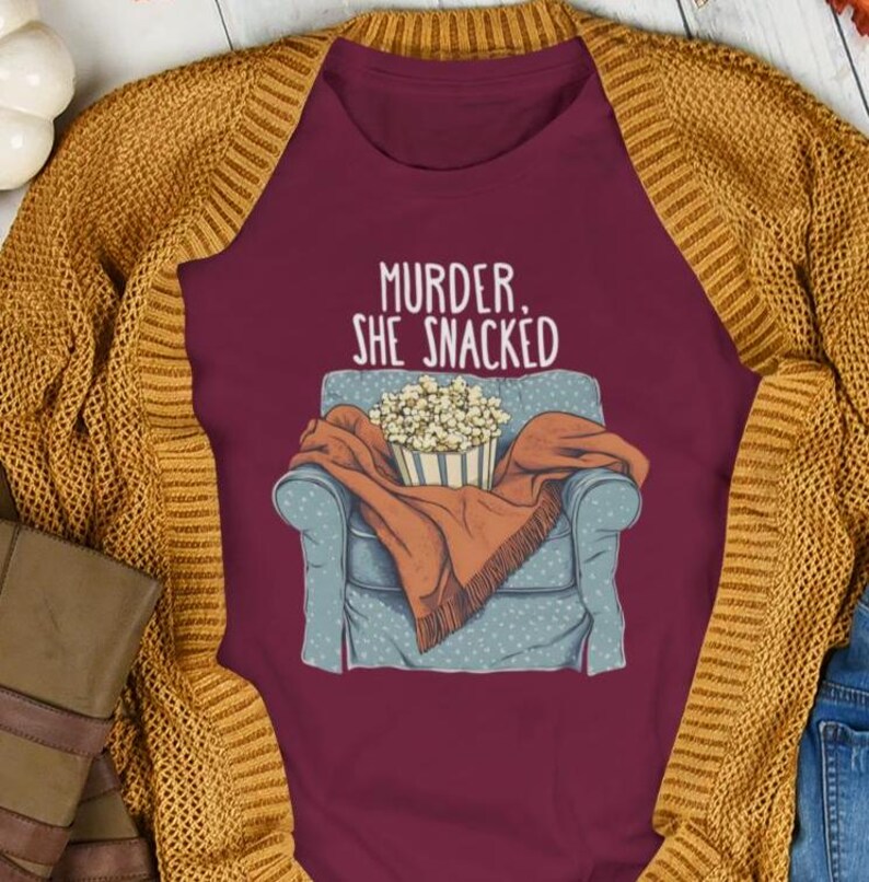 Op de afbeelding: Een maroon t-shirt met een afbeelding van iemand die in een fauteuil zit met een deken en een kom popcorn. De tekst "Murder, She Snacked" staat op het shirt gedrukt.