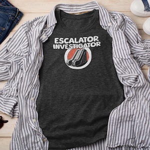 May include: Dark gray t-shirt with the words "ESCALATOR INVESTIGATOR" in white, above a graphic of an escalator inside a red circle. The shirt is laid on a striped button-down shirt and light wood surface.