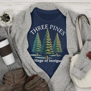 May include: A navy blue t-shirt with a white graphic of three pine trees and the text "Three Pines Village of Intrigue".