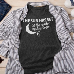 Op de afbeelding: Een donkergrijs T-shirt met de tekst "The sun has set let the murder mystery begin!" in wit gedrukt. Een halve maan en sterren zijn ook op het shirt gedrukt.