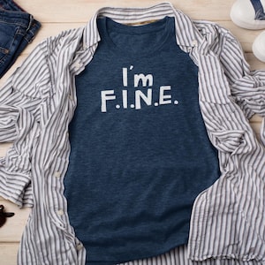 May include: A navy blue t-shirt with the white text "I'm F.I.N.E." is layered under a striped button-down shirt. The button-down shirt is open, revealing the t-shirt. The shirts are on a light wood surface.