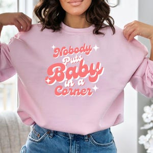 Peut inclure: Sweat-shirt rose avec le texte "Nobody Puts Baby in a Corner" dans une police rétro.