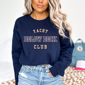 May include: Navy blue sweatshirt with the text "YACHT BELOW DECK CLUB" in gold lettering. The sweatshirt is a crew neck style and is being worn with blue jeans. The overall theme is nautical.