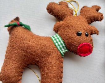 May include: A brown felt reindeer ornament with a green and white gingham ribbon around its neck. The reindeer has a red felt nose and black felt eyes. The ornament is hanging from a gold string.