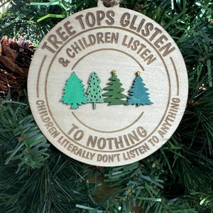 May include: A round wooden Christmas bauble with the text "Tree Tops Glisten & Children Listen To Nothing Children Literally Don't Listen To Anything". The bauble features three green stylised Christmas trees with gold stars.