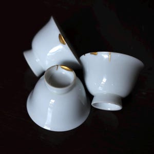 Może przedstawiać: Trzy białe filiżanki do herbaty z porcelany ze złotym akcentem na obrzeżu. Filiżanki są ułożone na ciemnej powierzchni, a światło odbija się od gładkiego, błyszczącego wykończenia.