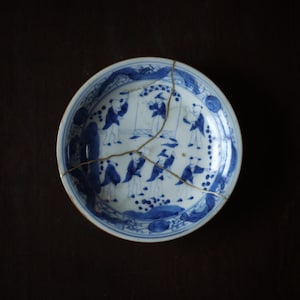 May include: A round ceramic plate featuring blue and white hand-painted illustrations of figures in a landscape. The plate has been repaired using the kintsugi technique, with visible gold-colored lacquer lines connecting the fractured pieces.