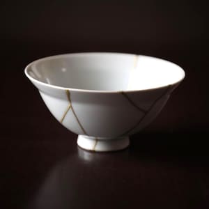 May include: A white ceramic bowl with a round base and a wide rim. The bowl has gold-colored lines that run across the surface, indicating the Kintsugi repair method. The bowl is set against a dark background.