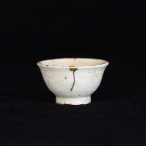 May include: A small, white ceramic bowl with a textured surface. The bowl has been repaired with gold-coloured kintsugi, a Japanese art form. The bowl is set against a black background.